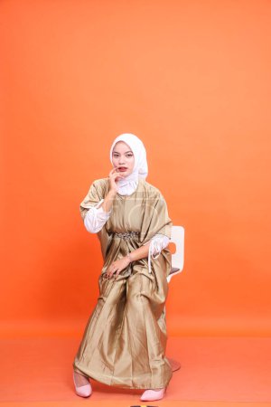 Full length studio portrait of young elegant Muslim Asian woman holding cheeks wearing kaftan hijab, high heels sitting on chair isolated orange background. Fashion, lifestyle, advertising concept