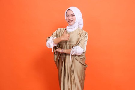 Beautiful cheerful indonesia girl model with her arms crossed with a thumbs up sign (okay good) wearing a kaftan Muslim dress with a hijab, isolated on an orange background. Concept lifestyle,fashion