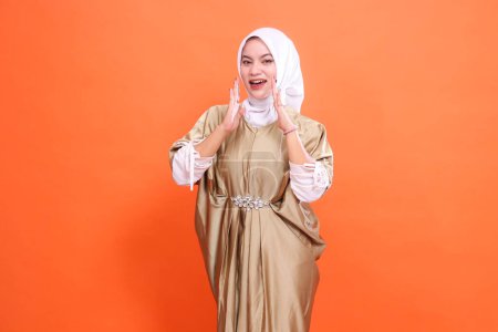 Cheerful beautiful young Asian woman model both hands announcing wearing Muslim dress kaftan with hijab, isolated on orange background. Concept of lifestyle, stylish fashion, promotion