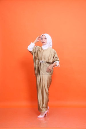 Full body of young elegant indonesia Muslim woman wearing kaftan hijab standing sideways left holding head and waist isolated orange background. The concept of Islamic religion, fashion and commercial