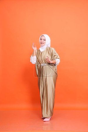 Full body of young cheerful Asian Muslim woman wearing kaftan hijab standing happy pose isolated orange background. Concept of Islamic religion, fashion and advertising