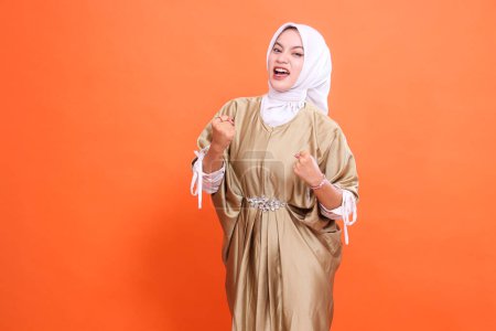 the expression of a beautiful Asian girl, cheerful, emotional, hands clenched in front of her, wearing a kaftan Muslim dress with a hijab, isolated on an orange background. Fashion, promotion concept