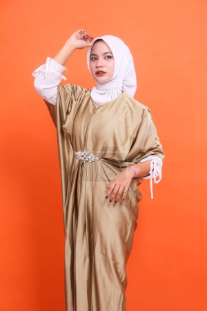 Young indonesia Muslim woman in hijab isolated orange background. standing elegantly, hands holding head to the side, candid wearing Islamic kaftan clothing. Half body portrait for religious, fashion