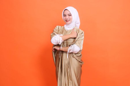 Beautiful cheerful Asian woman model with her arms crossed with a thumbs up sign (okay good) wearing a kaftan Muslim dress with a hijab, isolated on an orange background.stylish fashion, promotion