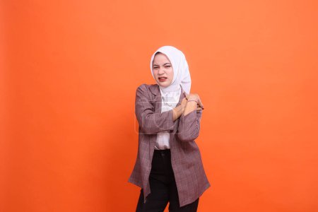 Porträt der schönen jungen indonesischen Frau, die sehr krank im Hijab offen ist, Erfahrungen Schulterschmerzen Rückenschmerzen Schwäche, legt Hand auf Schulter mit orangefarbenem Studiohintergrund. Gesundheitsversorgung