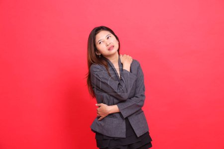 Expression de fille de bureau asiatique dans la douleur douleur d'épaule franche portant une veste grise et une mini-jupe noire sur fond rouge. pour concepts de santé, d'affaires et de publicité