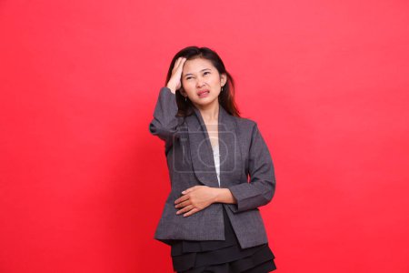 expresión de la mujer oficina indonesia sosteniendo su cabeza mareada con franqueza usando una chaqueta gris y falda sobre un fondo rojo. para conceptos de salud, negocios y publicidad
