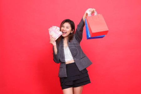 indonesia office woman enjoys looking at the camera carrying rupiah money and holding up various shopping paper bags wearing a gray suit on a red background. for transaction, lifestyle and business