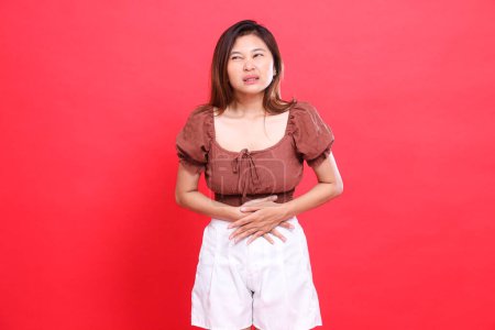 Indonesia cándida expresión de la mujer en el dolor con problemas estomacales con una blusa marrón y pantalones cortos sobre un fondo rojo. para conceptos de salud, estilo de vida y publicidad