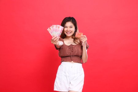 Cheerful Asian girl holding rupiah money in front of her and a debit credit card wearing a brown blouse with a red background. for transaction, technology and advertising concepts