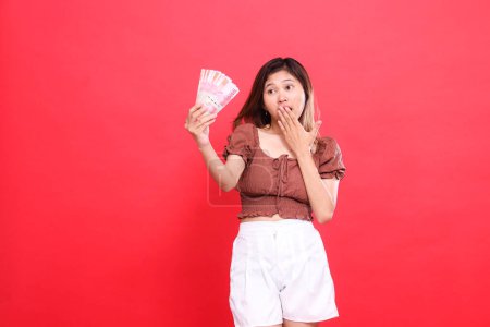The expression of a young indonesia woman is shocked, covering her mouth and holding rupiah bills up, wearing a brown blouse on a red background. for transaction, technology and advertising concepts