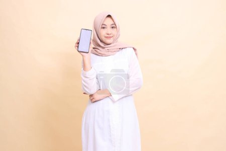 Photo for Woman asian muslim wearing a hijab, smiling, holding a cellphone gadget in her hand, arms crossed. Lifestyle, business and finance concept - Royalty Free Image