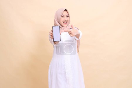 Photo for Asian Muslim woman wearing a hijab smiling happily holding a cell phone gadget in her hand while pointing at it. Lifestyle, business and finance concept - Royalty Free Image