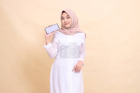 Photo for Young Asian Muslim woman wearing a hijab smiles holding a cellphone gadget slightly tilted in her hand. Lifestyle, business and commercial concept - Royalty Free Image