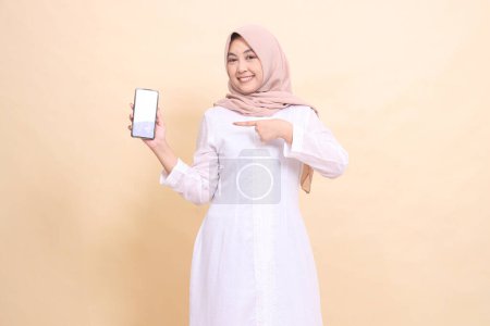 Photo for Young woman indonesia muslim wearing a hijab smiles cheerfully holding a smartphone gadget to the right in her hand while pointing at it. Lifestyle, business and commercial concept - Royalty Free Image