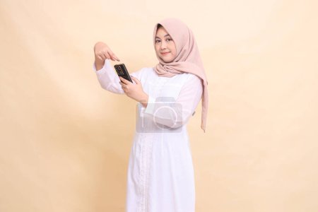 Photo for Asian Muslim woman wearing a hijab smiles at the camera, operating a cellphone gadget with both hands while pointing. Lifestyle, technology and promotion concept - Royalty Free Image