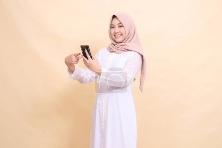 Photo for Asian Muslim woman wearing a hijab smiles cheerfully at the camera holding a cell phone gadget in her hand while pointing at it. Lifestyle, technology and promotion concept - Royalty Free Image