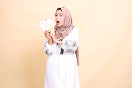 Foto de Retrato de una hermosa mujer musulmana asiática vestida con un hiyab, conmocionada y confundida, sosteniendo y mostrando un regalo o fitrah a la derecha en el Eid. utilizado para la publicidad, regalos, Eid y Ramadán - Imagen libre de derechos
