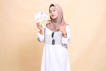 Porträt einer schönen asiatischen Muslimin, die einen Hijab trägt, anmutig lächelt, eine Daumen-nach-oben-Geste macht und am Eid-Tag ein Geschenk oder eine Fitra nach rechts hält. verwendet für Werbung, Eid und Ramadan
