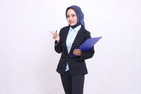 Asian office woman wearing a hijab standing smiling cheerfully pointing right at the camera carrying a pen and hugging a clipboard. Beautiful Muslim women wearing blue shirts for business finance