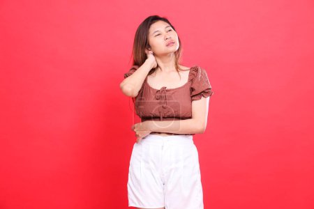 L'expression d'une femme indonésienne avec les bras croisés, tenant l'arrière de son cou dans la douleur, portant un chemisier brun et un short avec un fond rouge. concept de santé, de soins et de publicité
