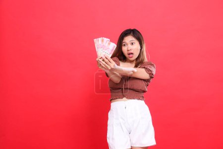 geste d'une jeune femme indonésienne choqué candide avec les deux mains tenant de l'argent rupiah devant elle portant un haut de chemisier brun avec un fond rouge. pour transaction, technologie et publicité