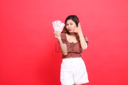 geste d'une jeune fille asiatique joyeuse tenant de l'argent rupiah devant et un panneau téléphonique. Je porte une blouse marron avec un fond rouge. pour les concepts de transaction, de technologie et de publicité