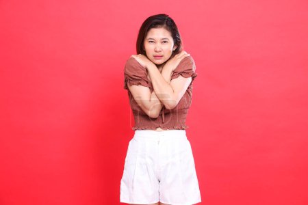 Der Gesichtsausdruck der Asiatin, offen, Angst, Körper, Schüttelfrost, Fieber, Erkältung, sie trägt eine braune Bluse und kurze Hosen auf rotem Hintergrund. für Gesundheits-, Lifestyle- und Werbekonzepte