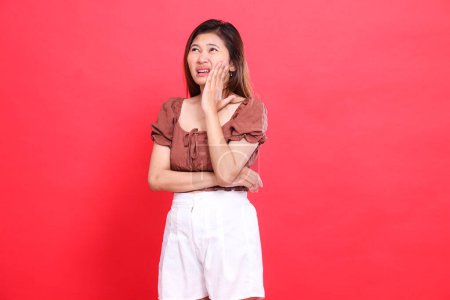 Die Geste der indonesischen Frau, offen, krumm nach rechts, hält ihre Wange, Zahnschmerzen, Schmerzen, trägt eine braune Bluse und kurze Hosen mit rotem Hintergrund. für Gesundheits-, Pflege- und Werbekonzept