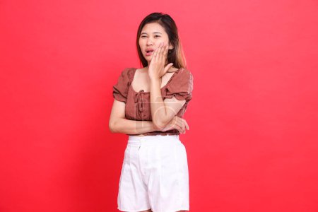 Geste einer Asiatin, die ihre Wange mit einem schmerzenden Zahn hält und eine braune Bluse und kurze Hosen mit rotem Hintergrund trägt. für Gesundheits-, Pflege- und Werbekonzept