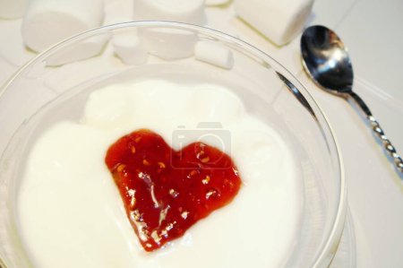 Foto de Postre rematado con mermelada de frambuesa en forma de corazón, cuchara retorcida de acero inoxidable y malvaviscos que caen sobre un plato blanco. - Imagen libre de derechos