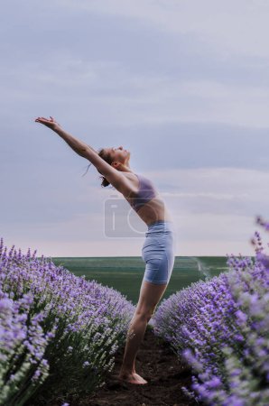 Mujer en ropa deportiva haciendo yoga pose backbend en medio de un campo de lavanda en un día nublado.