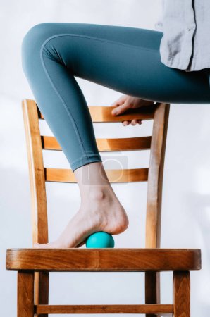 Frau macht Fußmassage mit Therapieball mittels Stuhlstütze