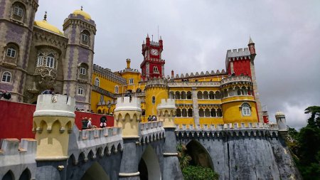 Photo for Sintra, Portugal - Jun 03, 2018: Exterior of Palacio Nacional da Pena, Palace Pena, with tourists walking under a cloudy sky, features the colourful walls of the palace before the pandemic - Royalty Free Image