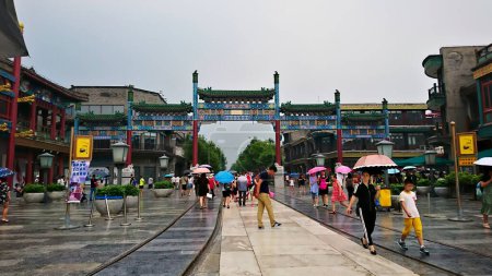 Foto de Qianmen Street, Beijing, China - 11 de agosto de 2018: Un colorido arco chino de Qianmen Street con turistas caminando por la calle bajo la lluvia y tiendas en los dos lados antes de la pandemia - Imagen libre de derechos