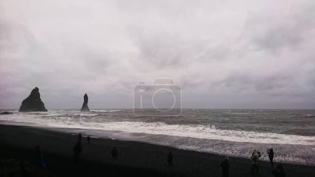 Photo for Reynisdrangar, Iceland - 03.26.2018: Tourists walking on a black sand beach on a rainy day with waves crashing on the coast - Royalty Free Image