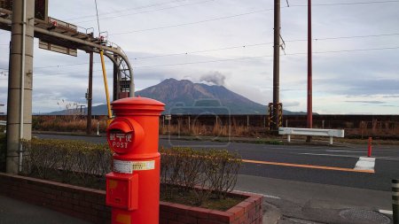 Foto de Kagoshima, Japón - 01.29.2020: Sakurajima libera humo bajo un cielo azul con un buzón rojo japonés en el frente en el pavimento a lo largo de una carretera vacía - Imagen libre de derechos