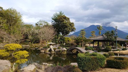 Photo for Kagoshima, Japan - 01.29.2020: A stone bridge over a pond with a pavilion on the side in Sengan-en and Sakurajima at the back releasing smoke under a cloudy sky - Royalty Free Image