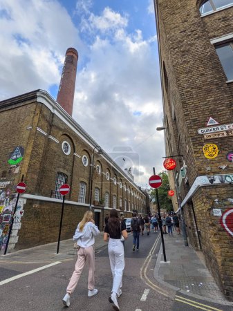 Photo for London, UK - 09.26.2021: Wide angle view of pedestrians walking on the busy Brick Land outside The Boiler House with a historic chimney under a cloudy blue sky - Royalty Free Image
