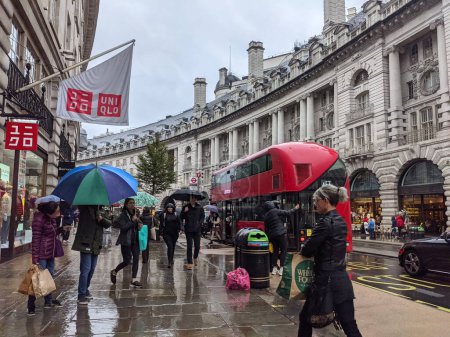 Foto de Londres, Reino Unido - 10.02.2021: Compradores con sombrillas caminando por Regent Street junto a Uniqlo con coches y un autobús de dos pisos desembarcando pasajeros en una parada de autobús bajo la lluvia - Imagen libre de derechos