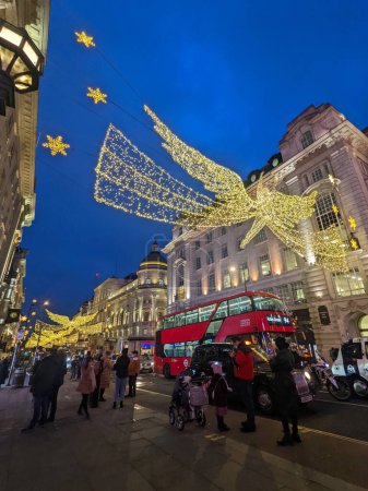 Foto de Londres, Reino Unido - 12.21.2021: Los peatones toman fotos del ángel Decoración de iluminación de Navidad colgada en Regent Street durante el crepúsculo con coches y un autobús de dos pisos que pasa - Imagen libre de derechos