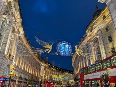 Foto de Londres, Reino Unido - 12.21.2021: Dos ángeles Iluminaciones de Navidad decoraciones colgando en la entrada de Regent Street con autobuses de dos pisos pasando py debajo de ella durante el crepúsculo - Imagen libre de derechos