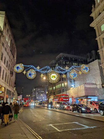 Photo for London, UK - 12.21.2021: Christmas lightings with stars hanging on Strand with pedestrians, double-decker buses, and cars passing under them at night - Royalty Free Image