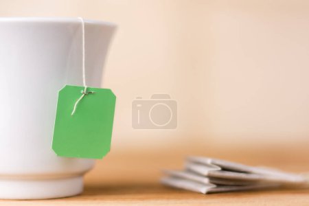 Teebeutel mit grünem Etikett in einer keramikweißen Tasse auf einer Tischplatte, warmgelber Hintergrund, Nahaufnahme einer Attrappe