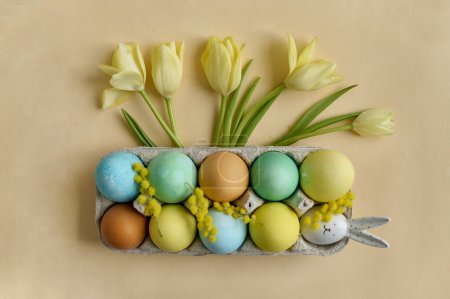 Foto de Colorful painted Easter eggs in a paper egg container with tulips on a beige background. Top view - Imagen libre de derechos