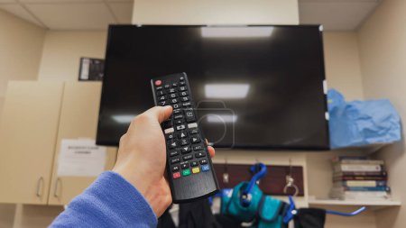 Photo for A person holding a remote control while sitting on a couch watching television. The sedentary lifestyle is countered by exercise equipment in the background, while the latest technology like smart TV - Royalty Free Image