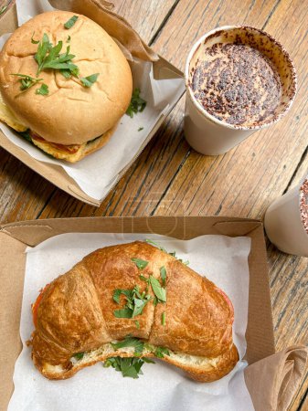 Photo for Top view of appetizing burger and croissant sandwich in cardboard boxes served on wooden table with disposable cups of foamy coffee in restaurant - Royalty Free Image