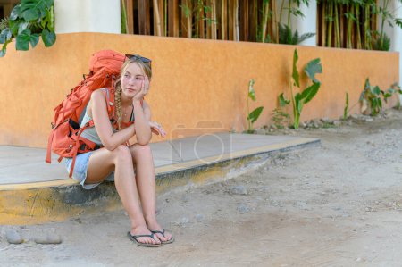 Téléchargez les photos : Cadre complet de jeune femme caucasienne aux longs cheveux blonds en vêtements décontractés avec sac à dos assis sur le trottoir et regardant loin en attendant le transport - en image libre de droit