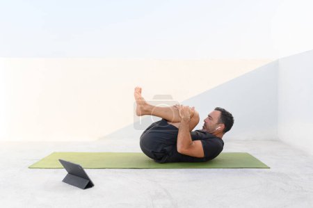 Photo for Middle aged man doing online yoga class alone on the terrace on sunny day wearing sport clothes earphones, ear bugs. High quality photo - Royalty Free Image