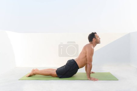 Téléchargez les photos : Homme d'âge moyen faisant de l'exercice à la maison, yoga étirement sur tapis sur une journée ensoleillée dans des vêtements sportifs. Photo de haute qualité - en image libre de droit
